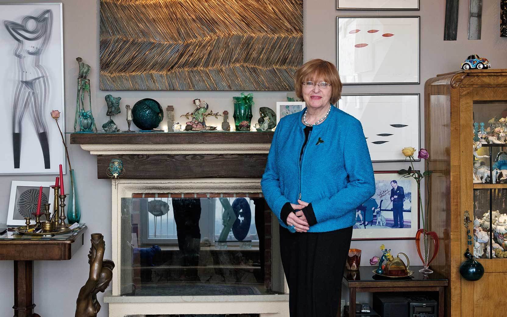 Older woman in a blue blazer stands smiling in an art-filled living room with a fireplace.