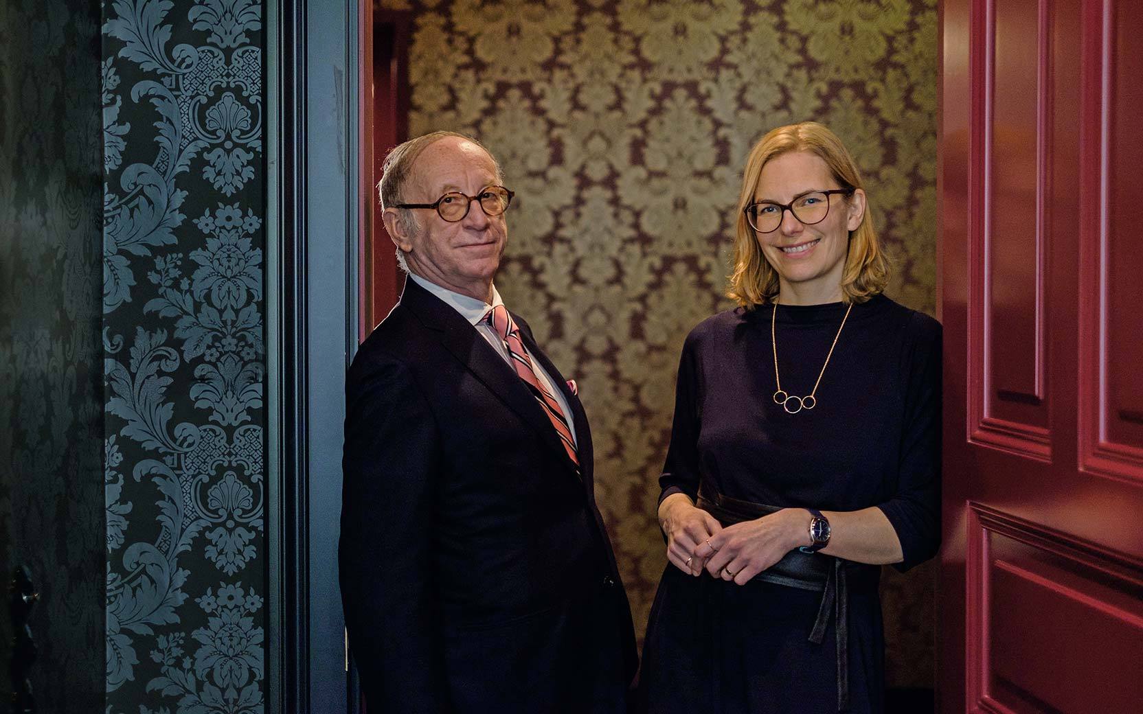 Two adults pose in a doorway with ornate wallpaper; man in a suit and pink striped tie, woman in a dark dress with necklace and glasses.