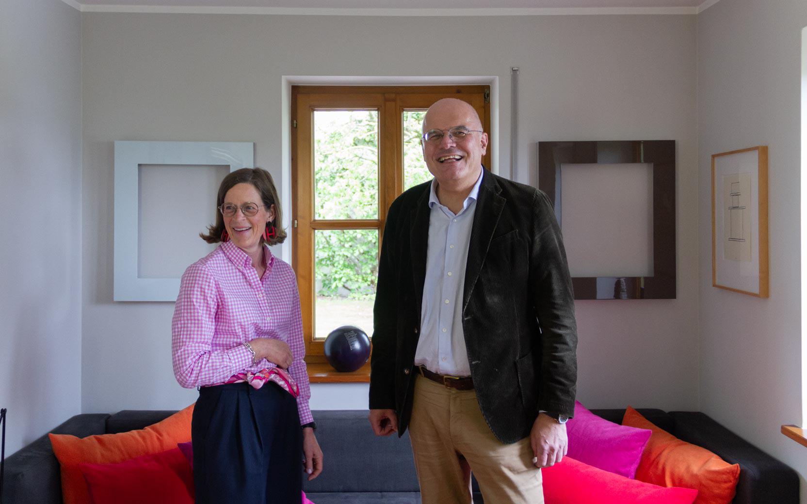 Two adults smiling in a living room; person in pink gingham shirt and navy skirt, other in blazer and khaki pants, sofa with orange cushions.
