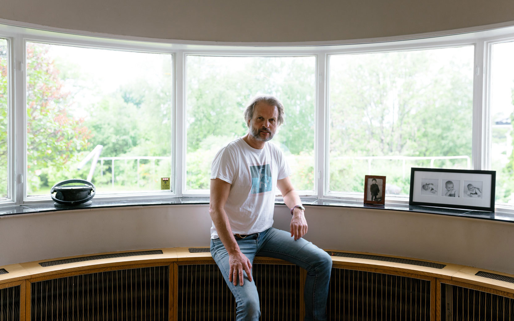 Adult with gray hair and beard sits on a curved bay window ledge, looking at the camera; framed photos and a garden outside.