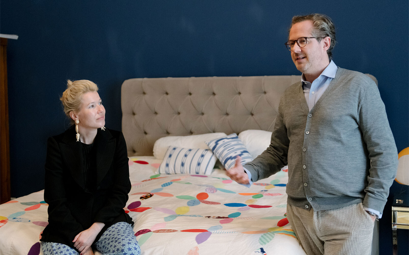 Adult wearing glasses gestures while speaking to seated adult on a colorful bedspread in a bedroom.