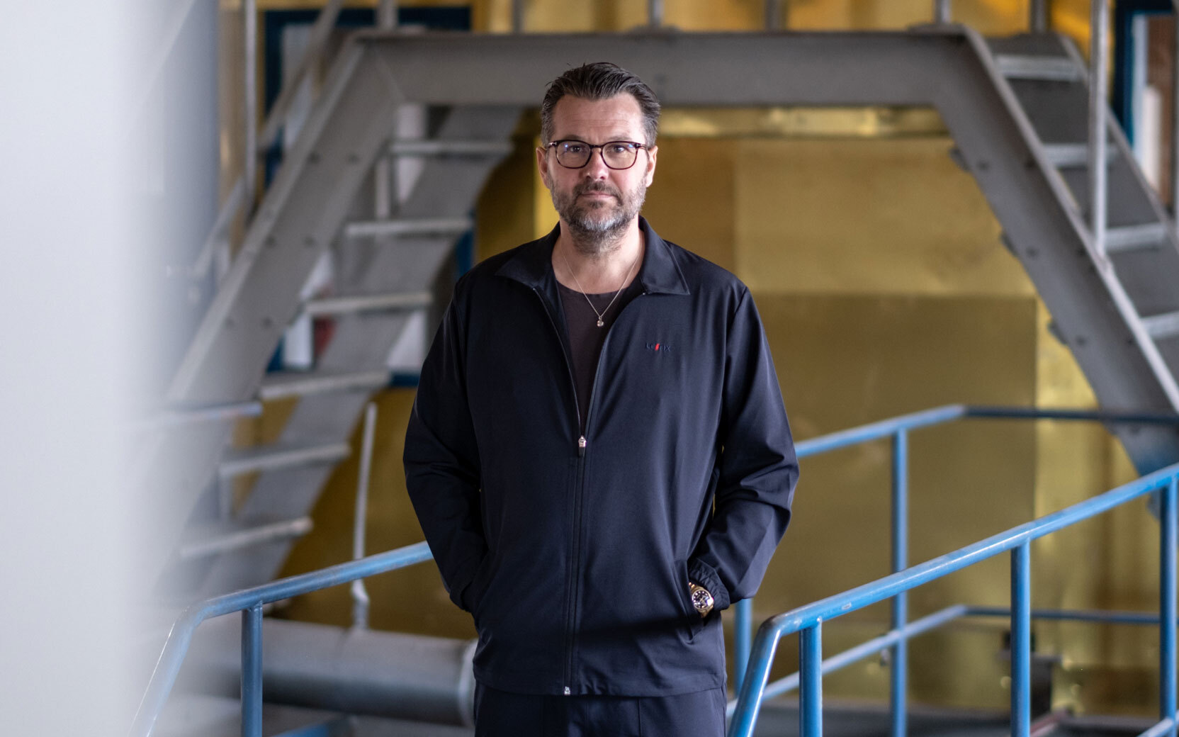 Person with glasses and beard wearing a black zip-up jacket stands with hands in pockets in an industrial setting with metal stairs and blue railings.