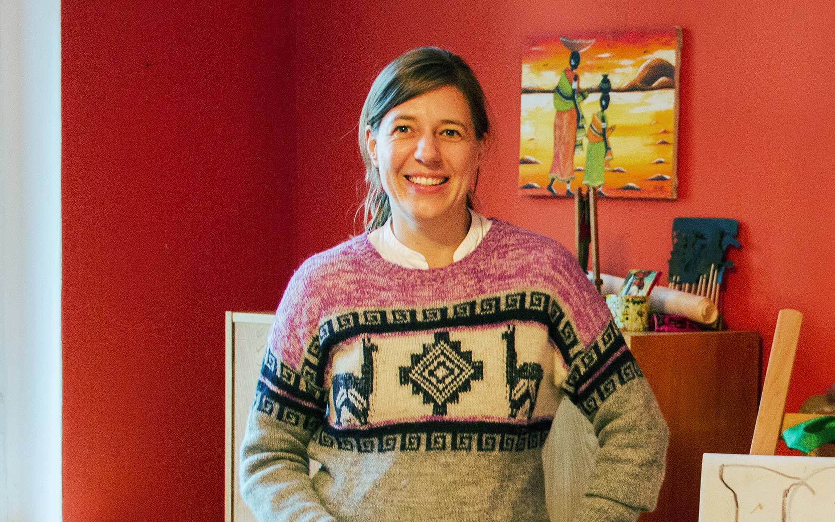 Lächelnde Person vor roter Wand, pink-grauer Pullover mit geometrischem Muster; Hintergrund mit Kunstwerk und Bastelmaterialien.