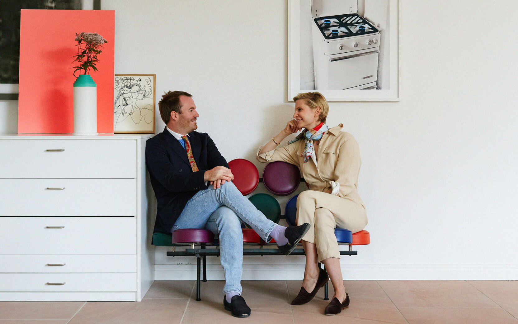 Two adults chat and smile on a colorful circular-seat bench in a bright modern room with white walls and art.