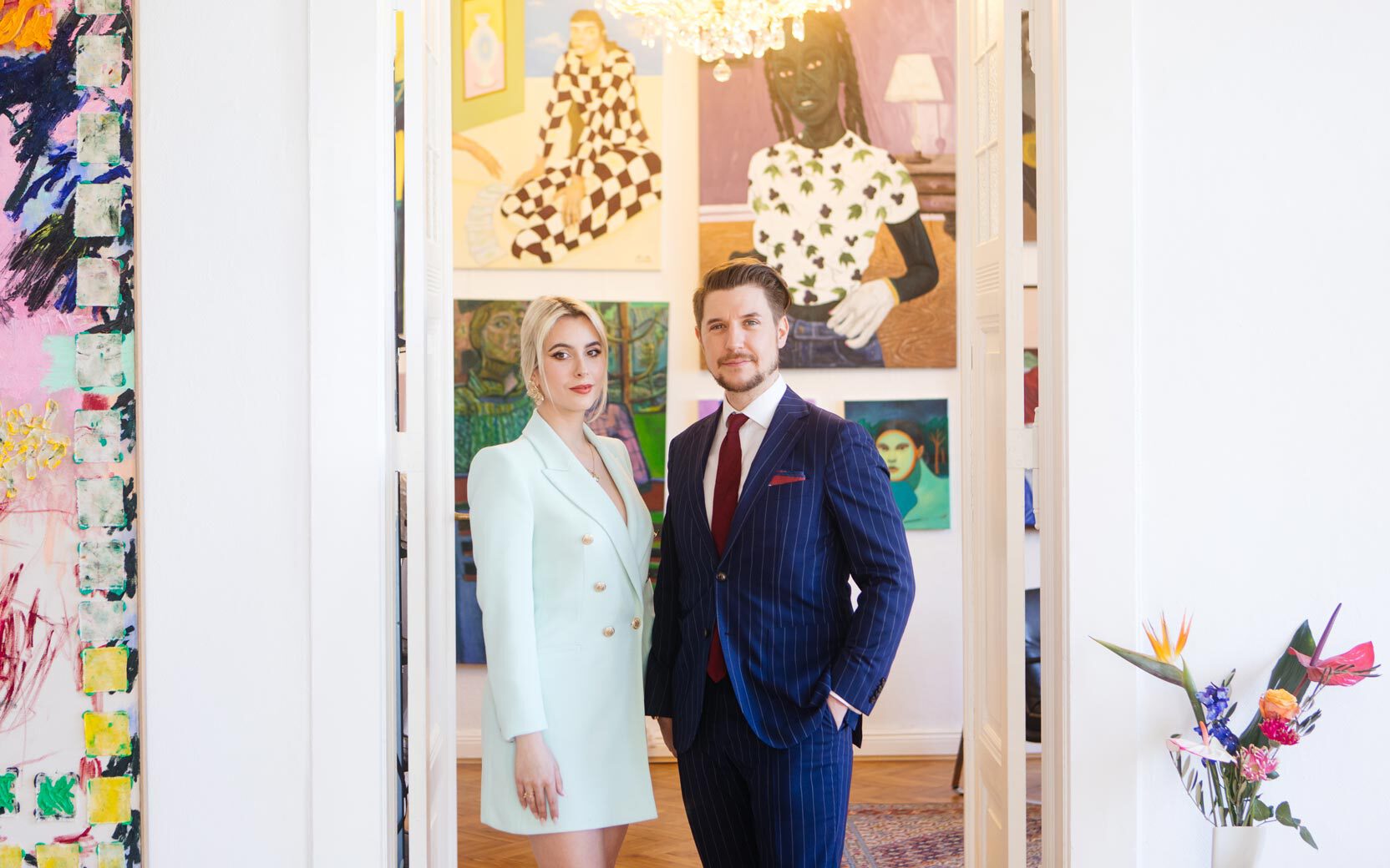 Two adults posing in an art gallery; woman in a mint blazer dress, man in a navy pinstripe suit, colorful artworks on the walls.