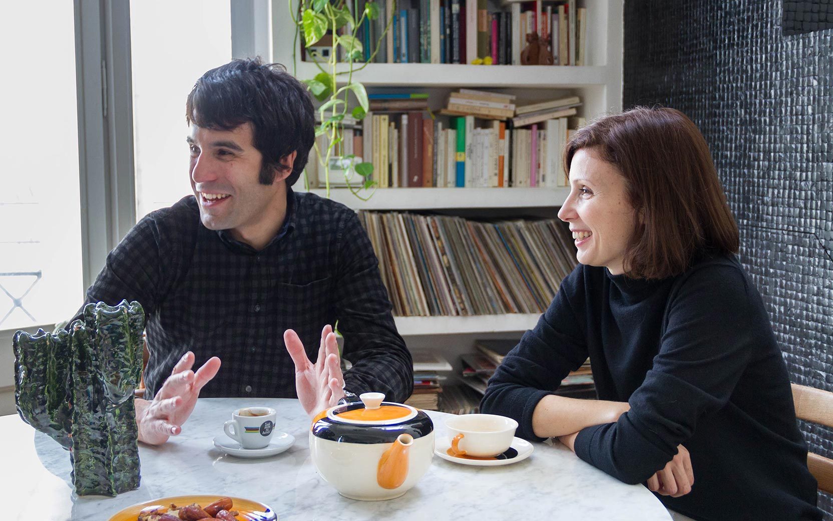 Two adults chat at a marble table in a bright room with a bookshelf; one gestures with hands, the other smiles. Teapot and cups on the table.