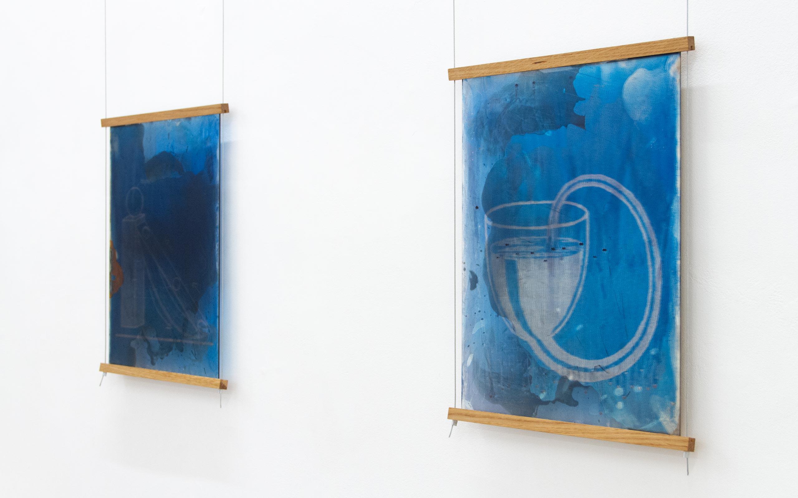 Two tall blue abstract panels hung on a white gallery wall, suspended by thin wires with wooden top and bottom bars.
