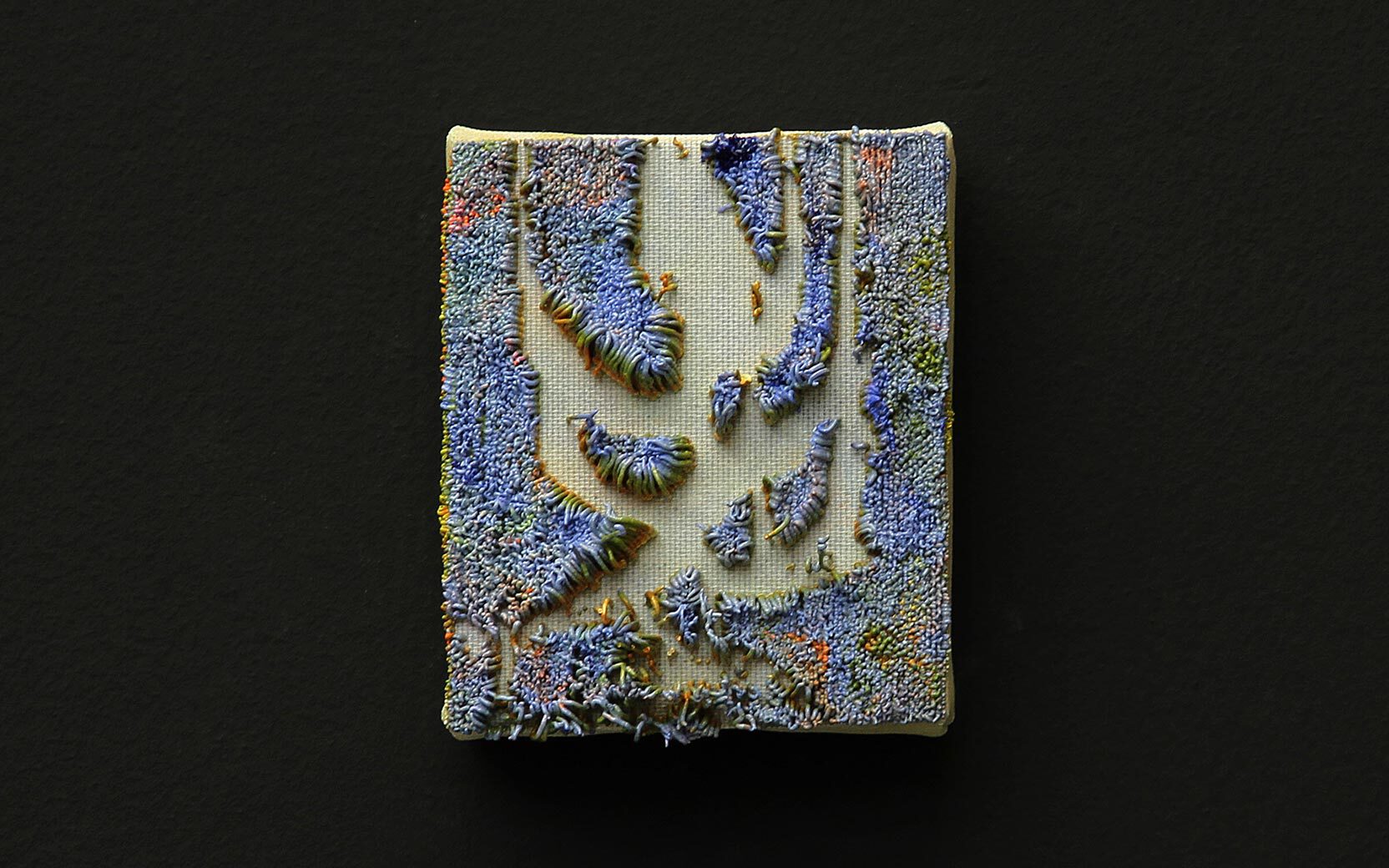 Small embroidered textile panel with raised blue stitches forming abstract shapes on beige fabric, set against a black background.