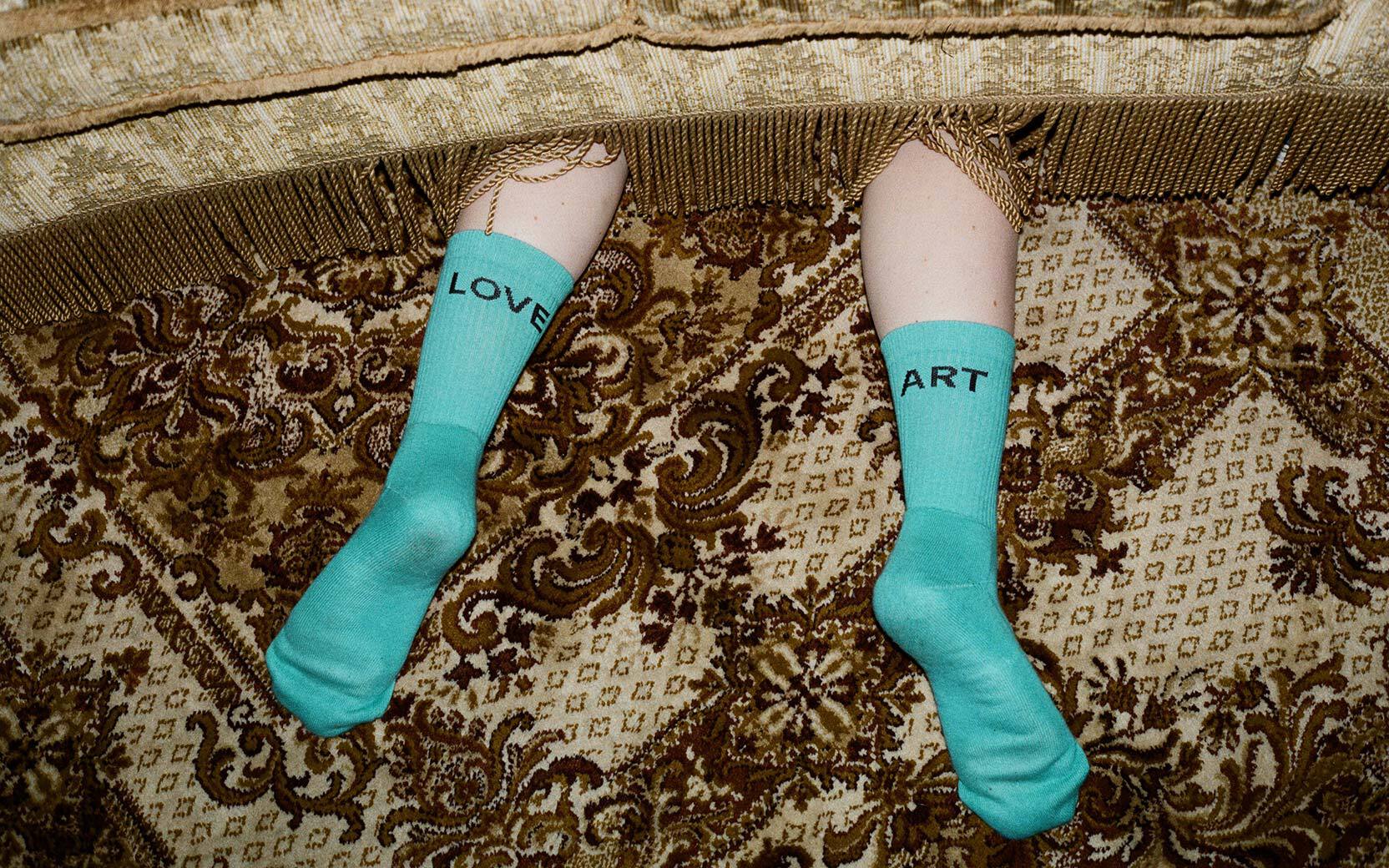 Lower legs in turquoise socks with LOVE and ART text, resting under a fringed sofa on a patterned rug.