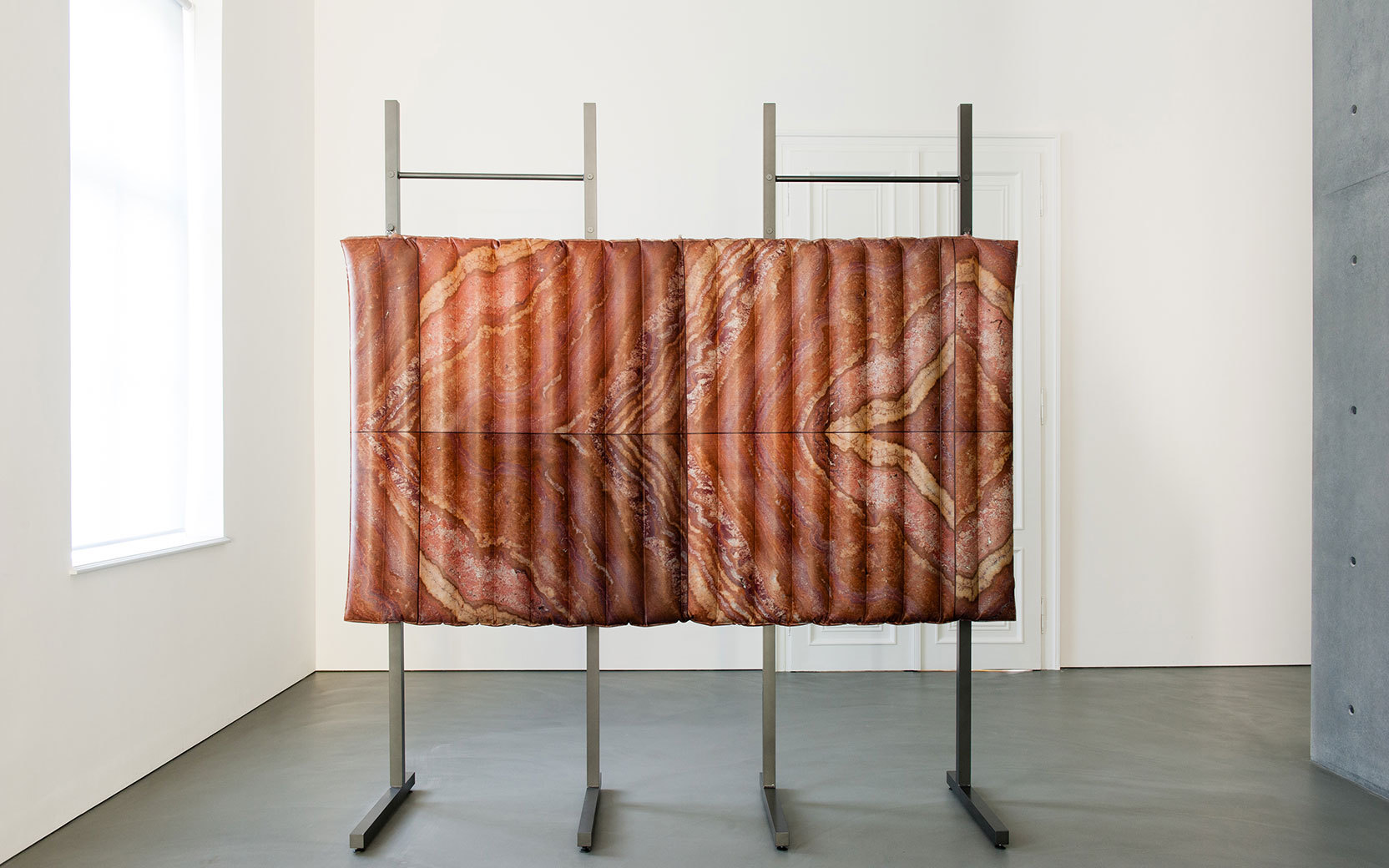 Kunstinstallation: gefaltete Stoffpaneele mit Fleischmuster, aufgehängt auf Metallgestell in hellem Ausstellungsraum.
