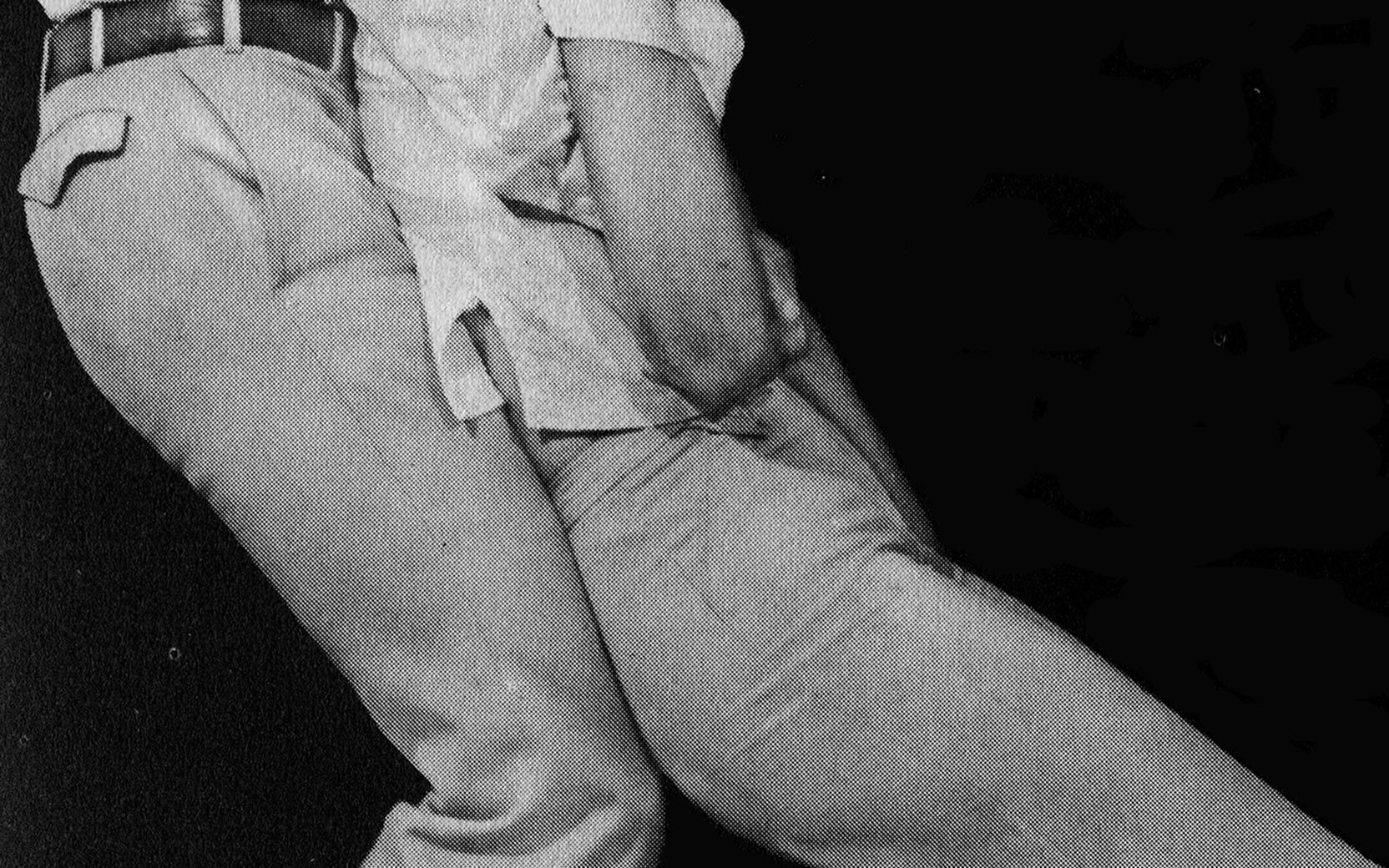 Person wearing rolled-up sleeves and belt sits, forearm resting on torso against a dark background