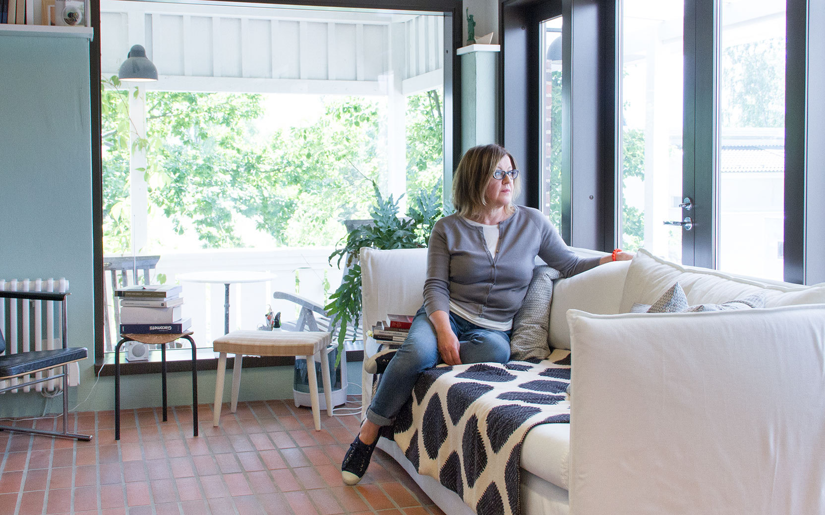 Person sitzt auf weißem Sofa im hellen Wohnzimmer, Blick nach rechts, Arm auf Lehne. Große Glas-Terrassentüren, Zimmerpflanzen, Bücherstapel daneben.