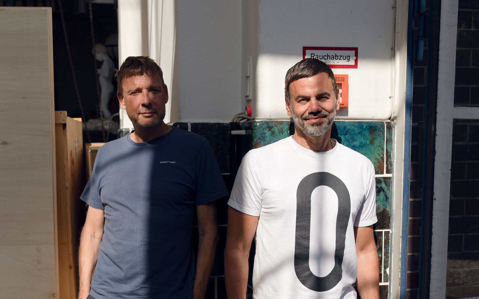 Two men stand side by side in a workshop area; one wears a blue t-shirt, the other a white t-shirt with a large black oval.