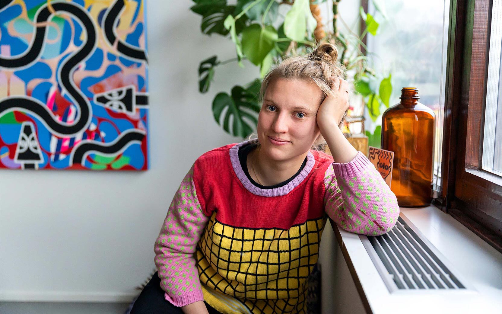 Person with blond hair in a bun rests their head on a hand by a windowsill with an amber bottle and green plants, beside a colorful abstract painting.