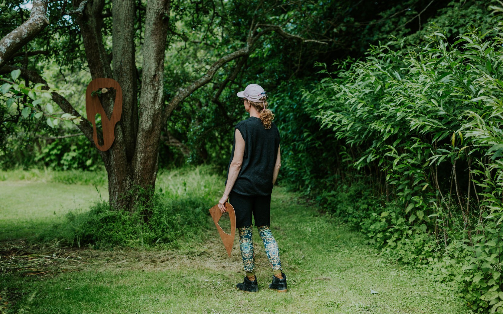 Person von hinten im Wald; pinke Mütze, dunkles Oberteil, bunte Leggings; hält hölzernes Herzloch und schaut Baumskulptur.