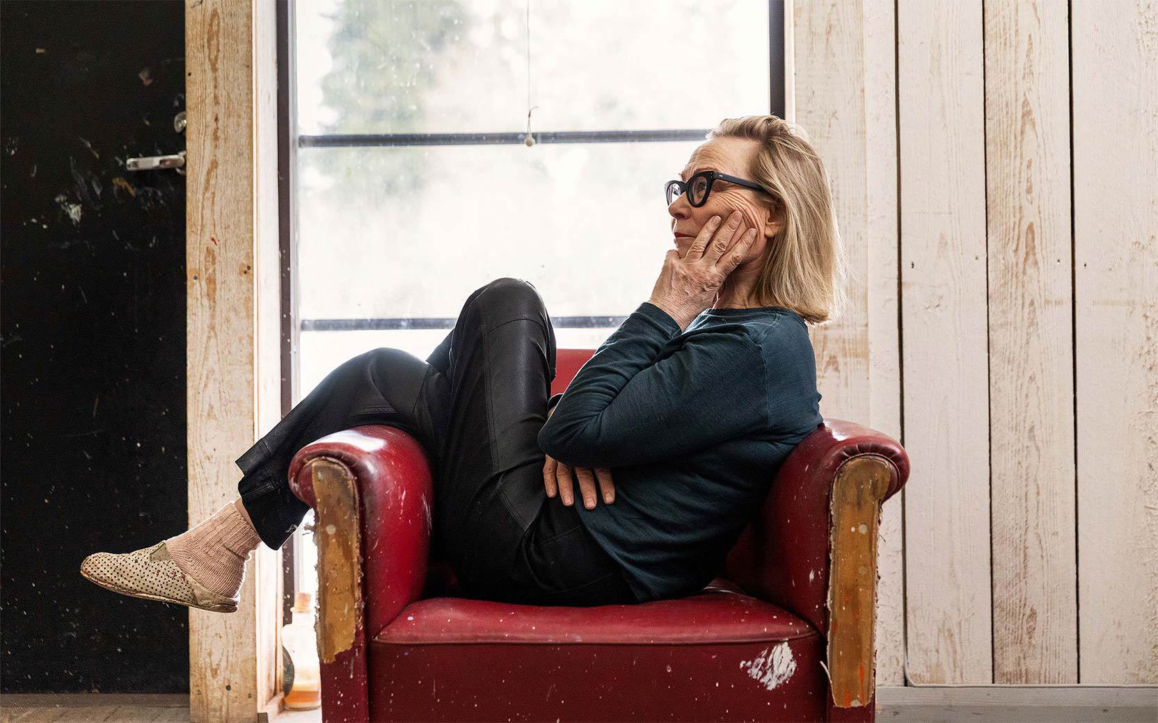 Older woman with glasses sits in a worn red armchair, hand on her cheek, legs drawn up, in a rustic room with wooden walls and a large window.