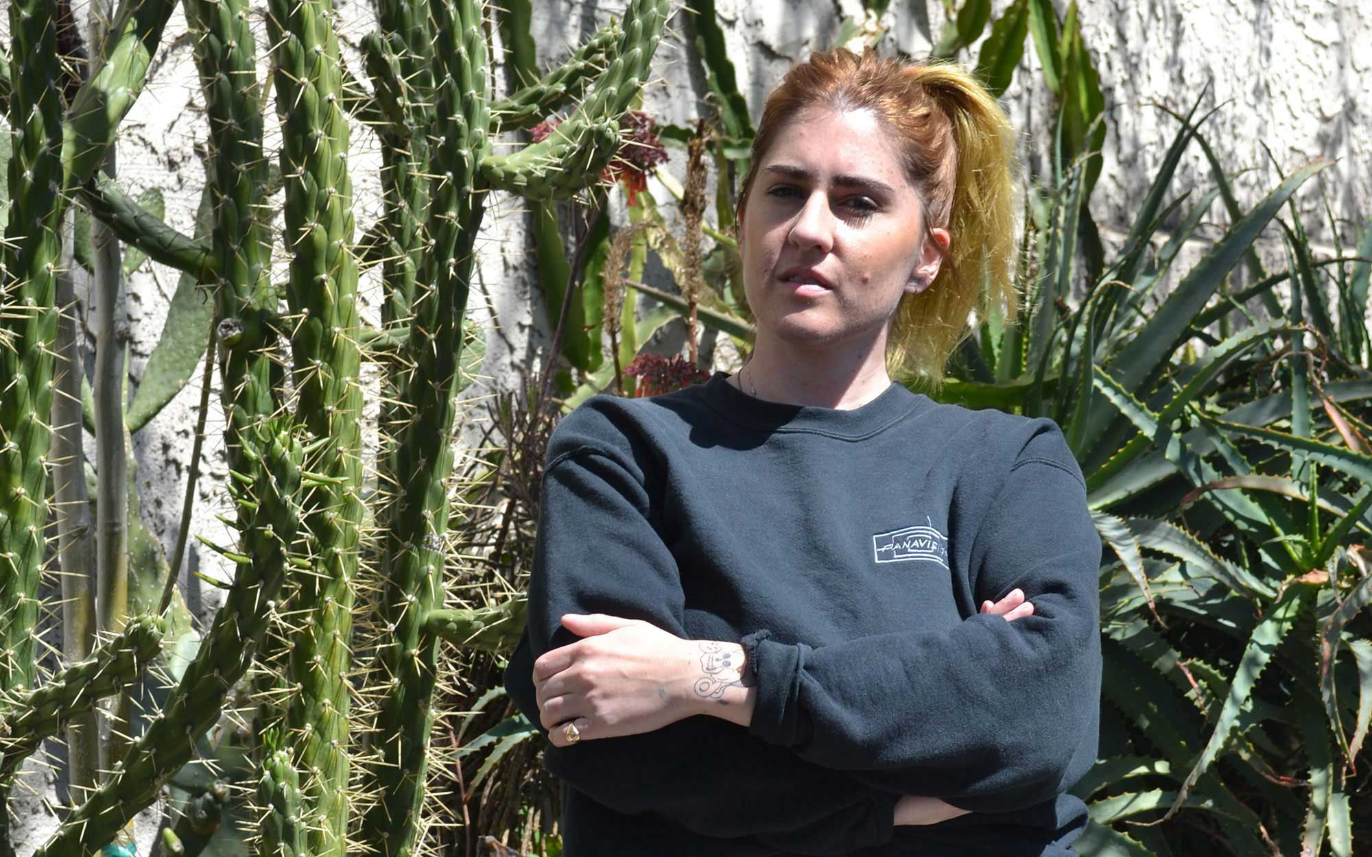 Frau mit hellbraunem Zopf, dunkles Sweatshirt, Arme verschränkt, vor Kakteen- und Sukkulentenpflanzen im Freien.