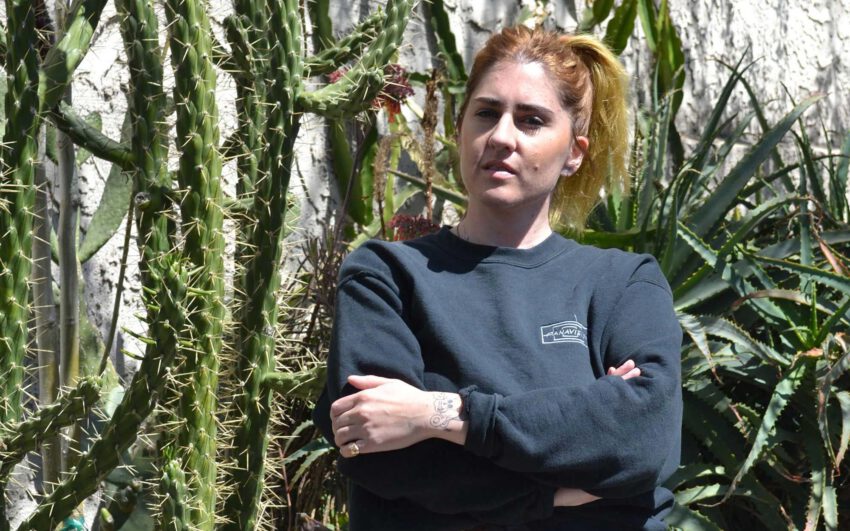 Person mit blondem Haar, dunkelblauem Sweatshirt, verschränkten Armen, vor Kakteen und Sukkulentenpflanzen im Freien