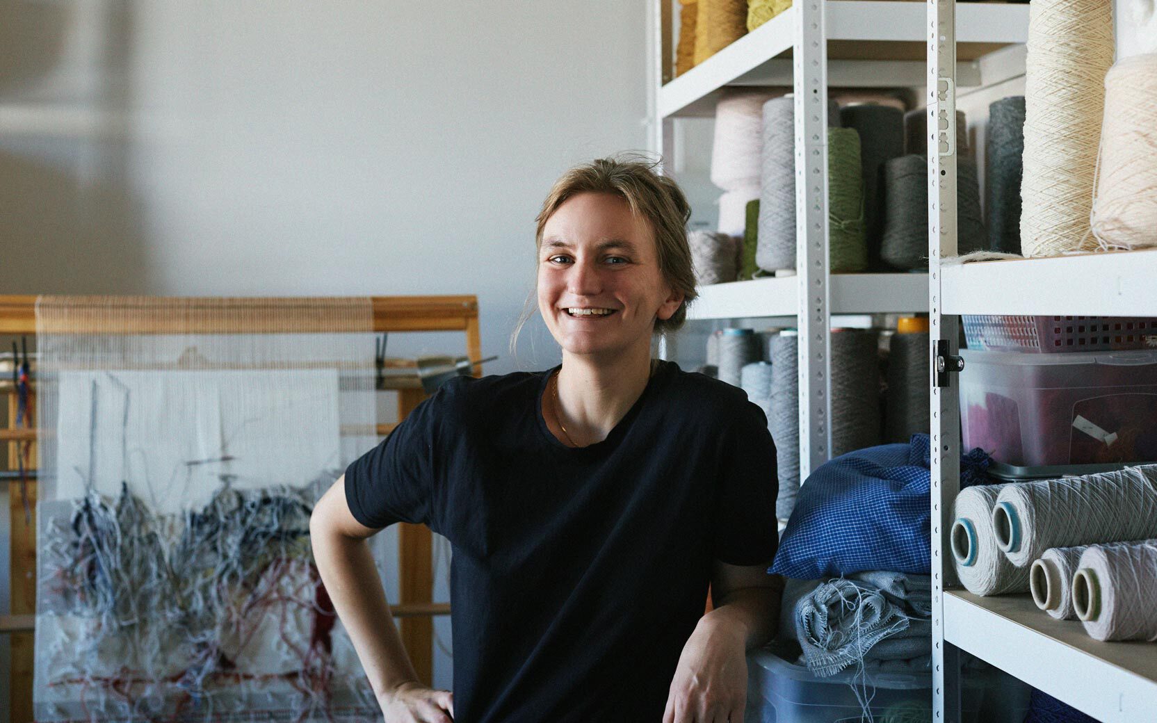 Smiling person in a black t-shirt stands in a textile workshop beside shelves of yarn cones and fabric rolls.