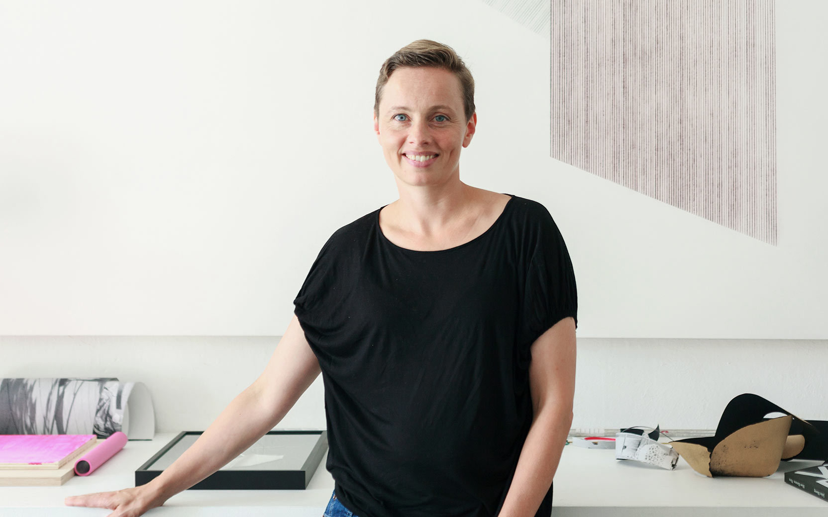 Lächelnde Frau in schwarzem T-Shirt steht an einem Atelier-Arbeitsplatz mit Papierrollen, Rahmen und Materialien.